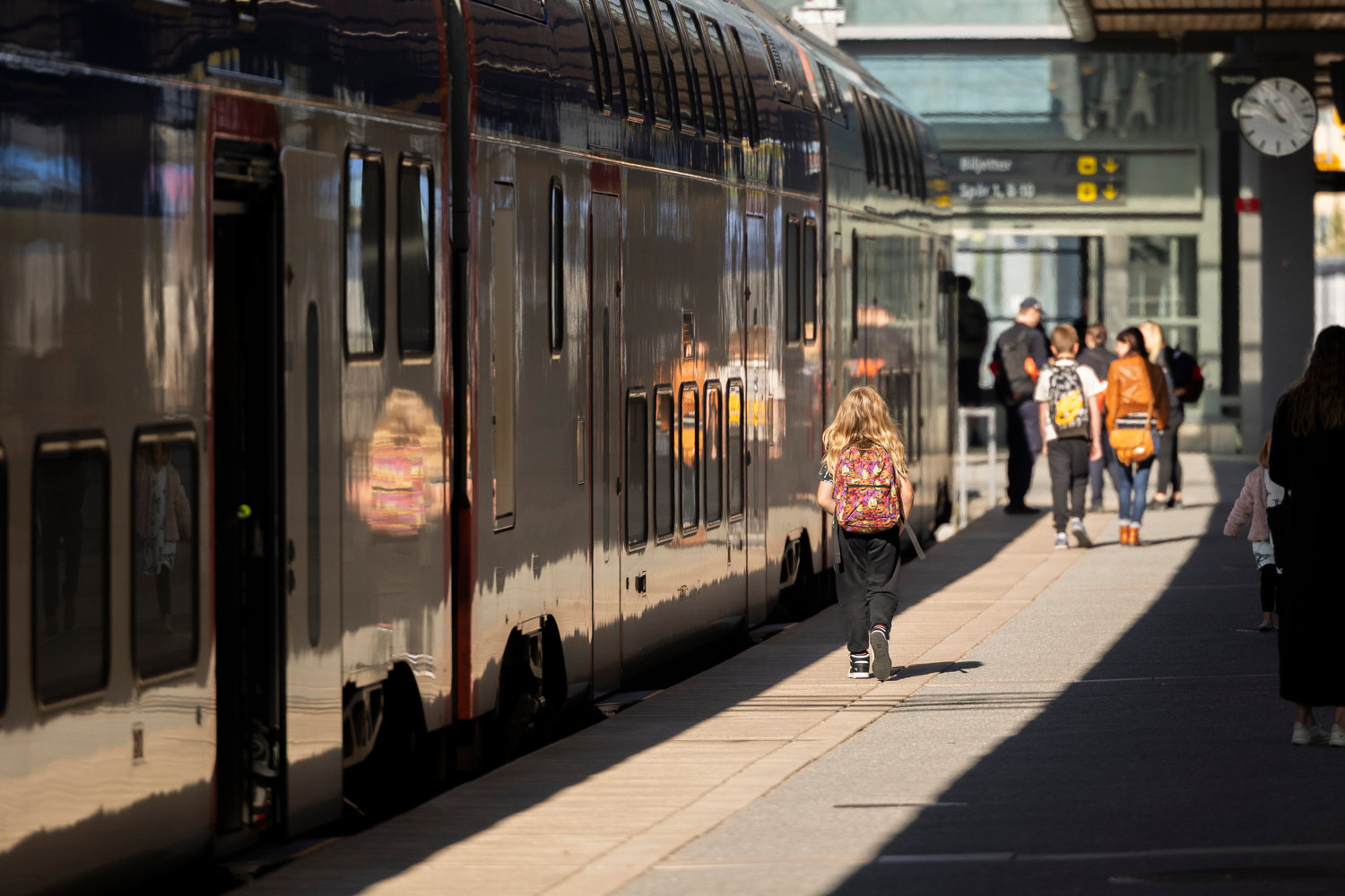 En grupp människor, inklusive barn med ryggsäckar, går längs en perrong bredvid ett stillastående tåg. Solen skiner och reflekteras på tågets yta