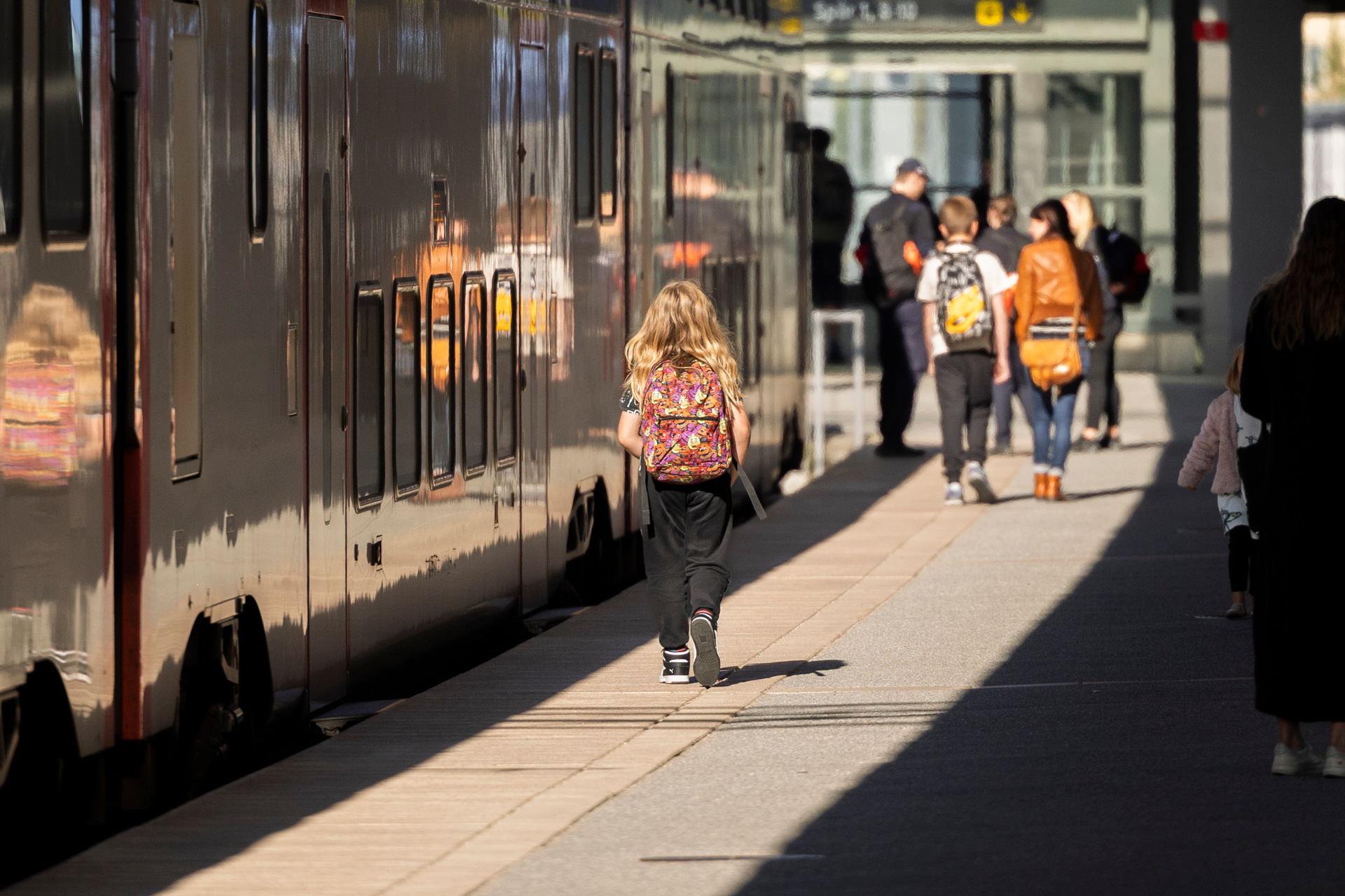 En grupp människor, inklusive barn med ryggsäckar, går längs en perrong bredvid ett stillastående tåg. Solen skiner och reflekteras på tågets yta